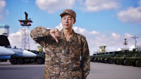 아시안 남자 군인 은 군 캠프 에 서 있는 동안 엄지손가락 을 아래 로 저지르며 카메라 를 향해 몸짓 을 하고 있다