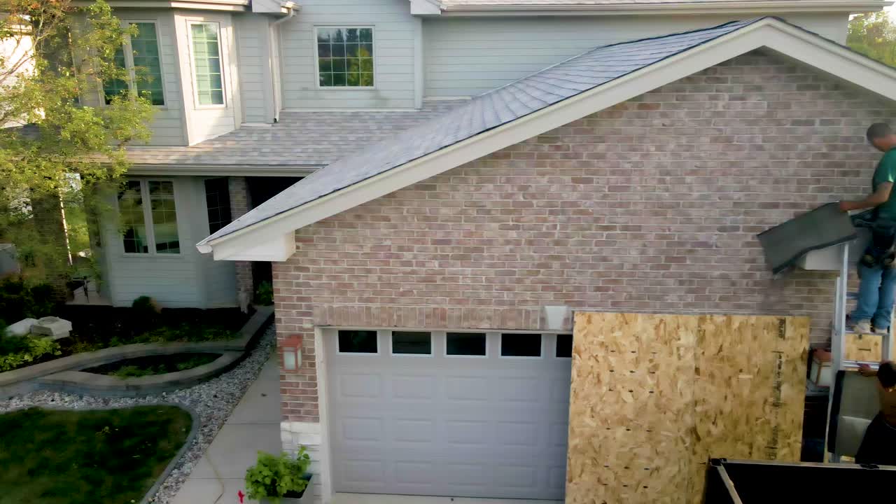 Construction worker repairing the exterior of a brick house, standing on a ladder and working on the roof area above the garage."