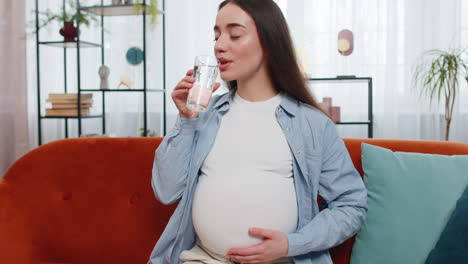 Pregnant-woman-holding-glass-with-water-and-drinking-while-sitting-on-sofa-healthy-maternity-concept