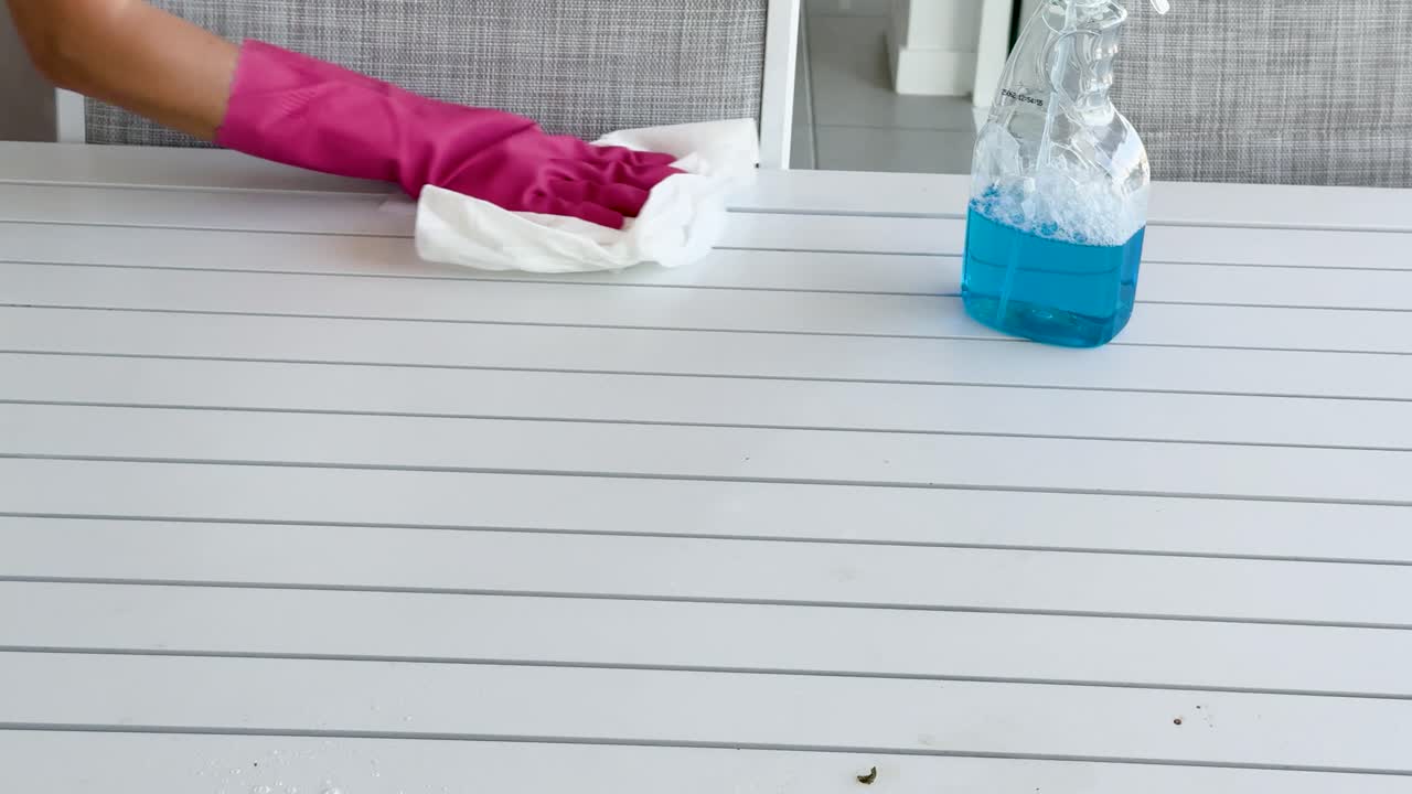 A person wearing pink gloves wipes a white table with a cloth and blue cleaning spray in a bright, modern indoor setting. Static camera, natural lighting