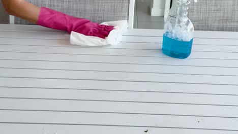 a person wearing pink gloves wipes a white table with a cloth and blue cleaning spray in a bright, modern indoor setting. static camera, natural lighting