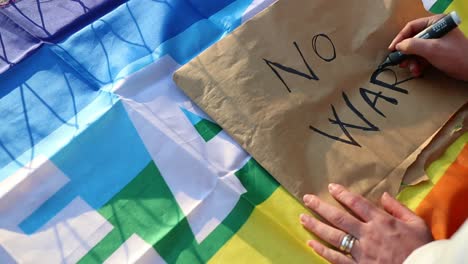 Female-caucasian-hand-writes-anti-war-protest-placard-with-a-peace-flag-as-background,-zoom-in-close-up-shot