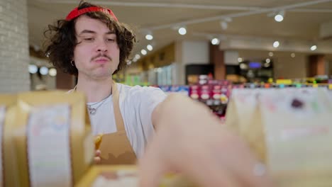 a brunette guy with curly hair in a cap chews a bun and takes inventory while rearranging goods on the shelves in a supermarket