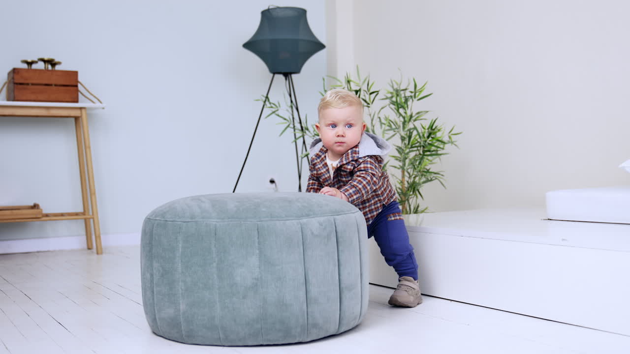 Adorable blond infant stands at the ottoman indoors. Baby looks around with surprise. Child learning to stand.