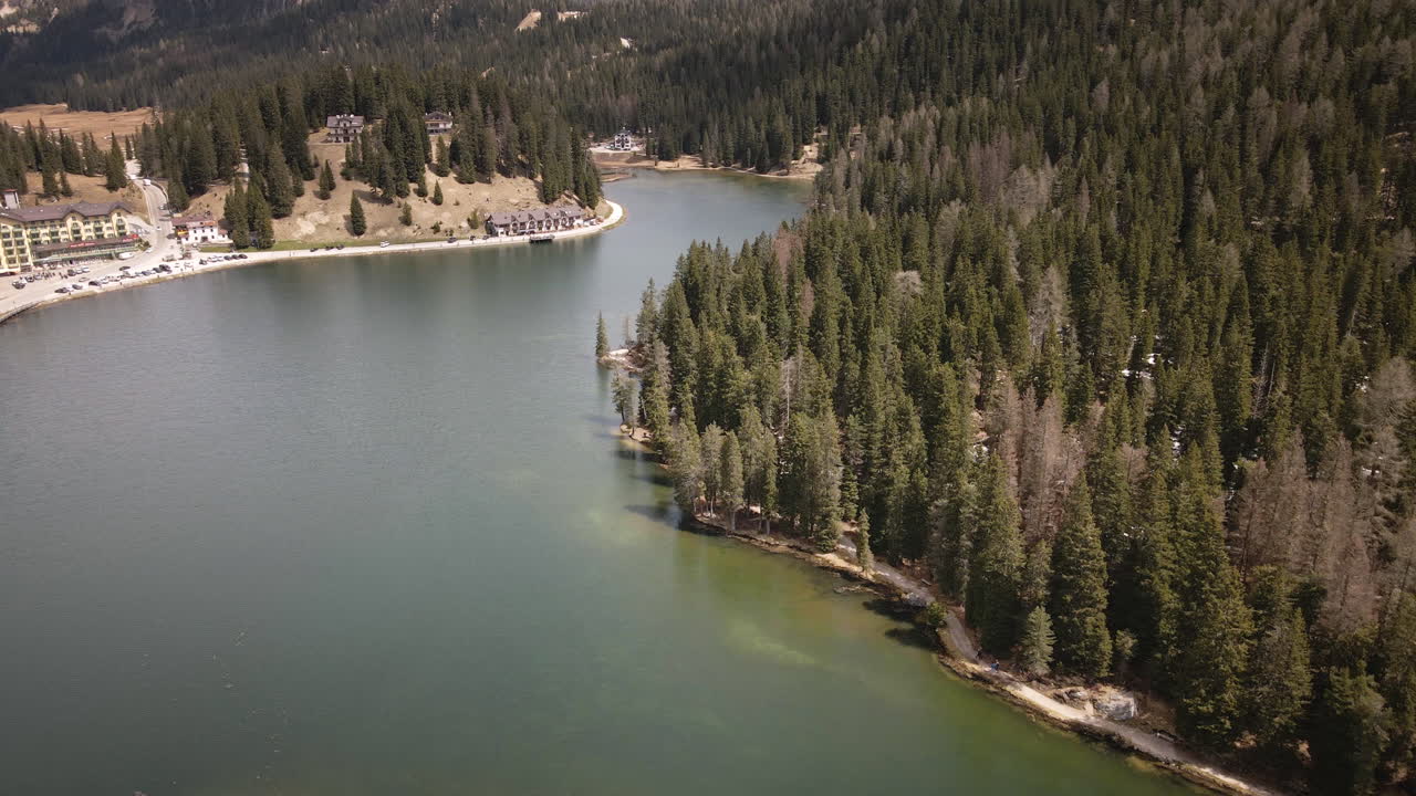 Camera moves forward high above a mountain lake, following the curve of the forested shoreline as alpine lodges and winding roads emerge beyond the dense evergreens