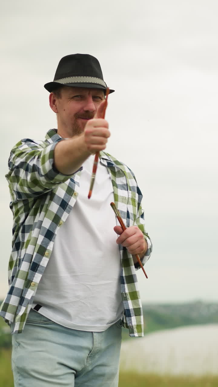 un pintor alegre con una camisa a cuadros y sosteniendo un pincel sonríe mientras una mujer borrosa es visible en el fondo. la escena captura un momento alegre y creativo en un entorno al aire libre