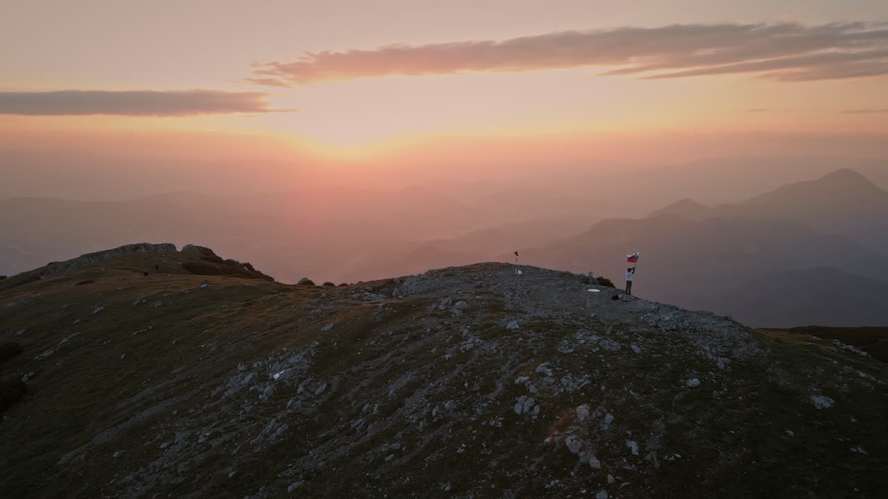 toma de drones de la cima de la montaña al amanecer, excursionista sosteniendo una bandera eslovena en el pol que está revoloteando en la brisa