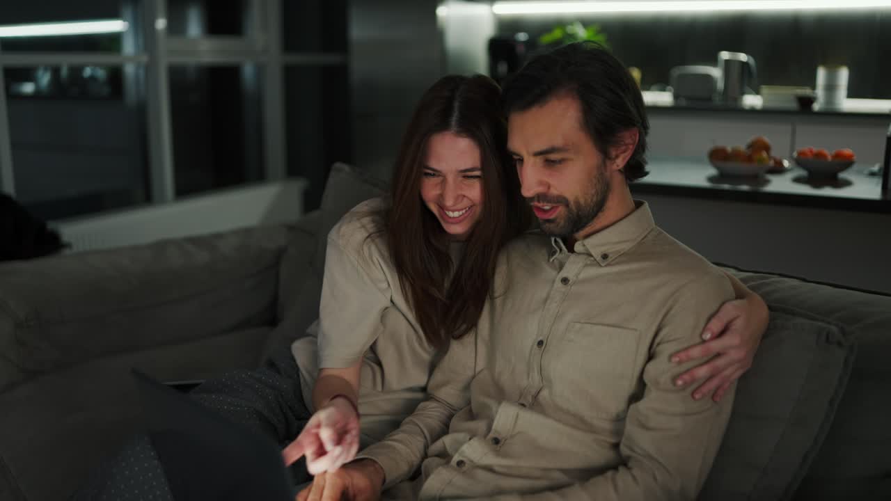 Happy brunette girl has fun and rejoices hugging her brunette husband with stubble in a beige shirt while he works remotely using a gray laptop on the sofa in a modern apartment in the evening