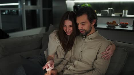 Happy-brunette-girl-has-fun-and-rejoices-hugging-her-brunette-husband-with-stubble-in-a-beige-shirt-while-he-works-remotely-using-a-gray-laptop-on-the-sofa-in-a-modern-apartment-in-the-evening