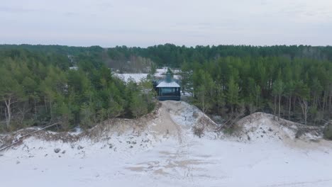 Aerial-establishing-view-of-Baltic-sea-coast-on-a-overcast-winter-day,-a-tiny-gray-holiday-house-at-the-beach-with-white-sand,-coastal-erosion,-climate-changes,-wide-drone-shot-moving-backward