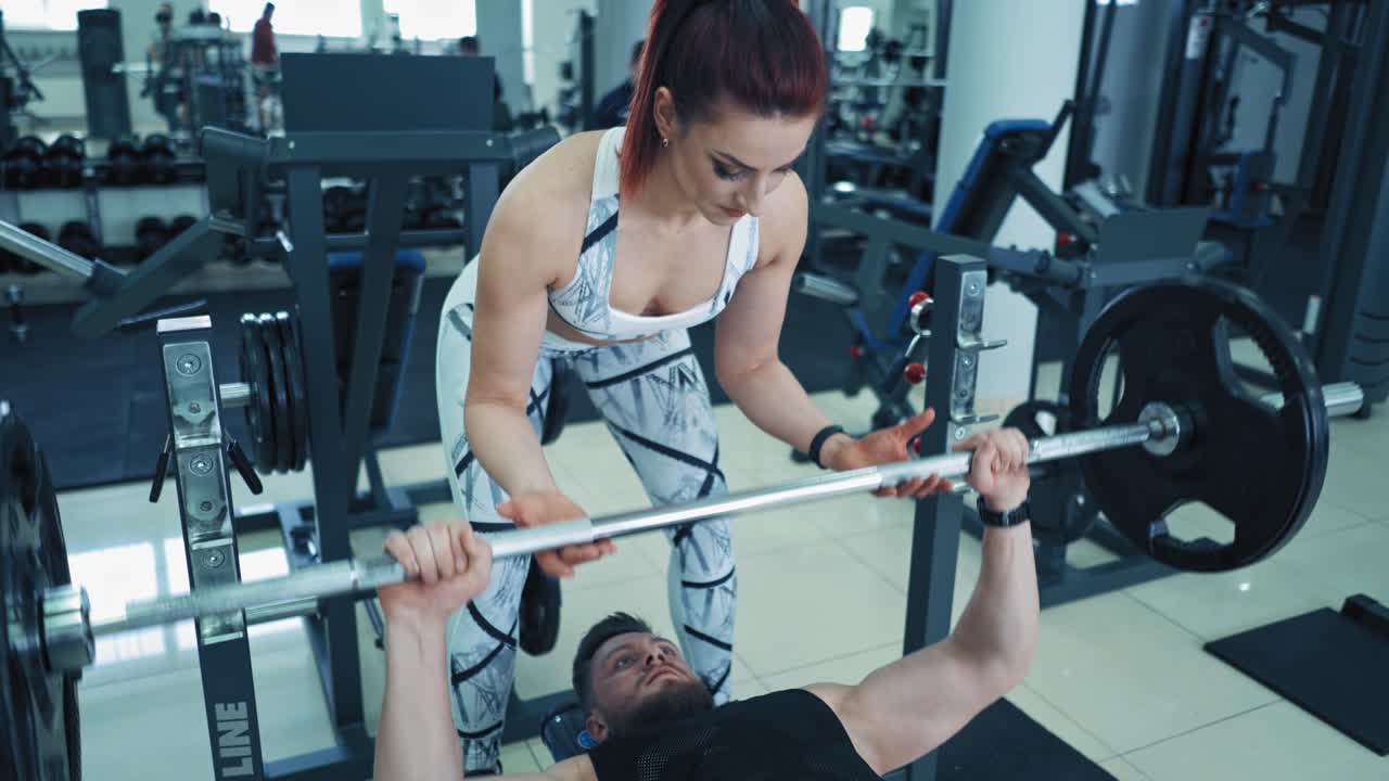 Strong bodybuilder lifting barbell in gym. Athletic man lying on special simulator and lifts barbell. Personal instructor woman helps athlete during training.