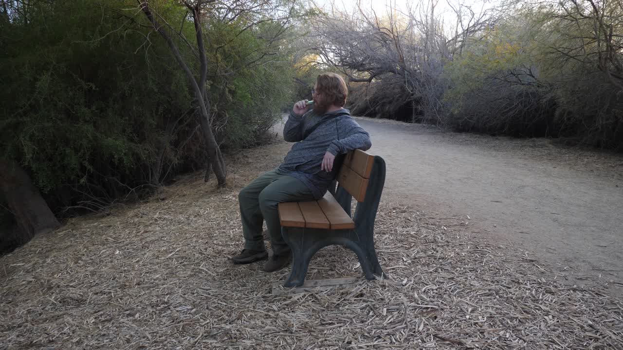 un hombre pelirrojo sentado y vaping en un banco del parque.