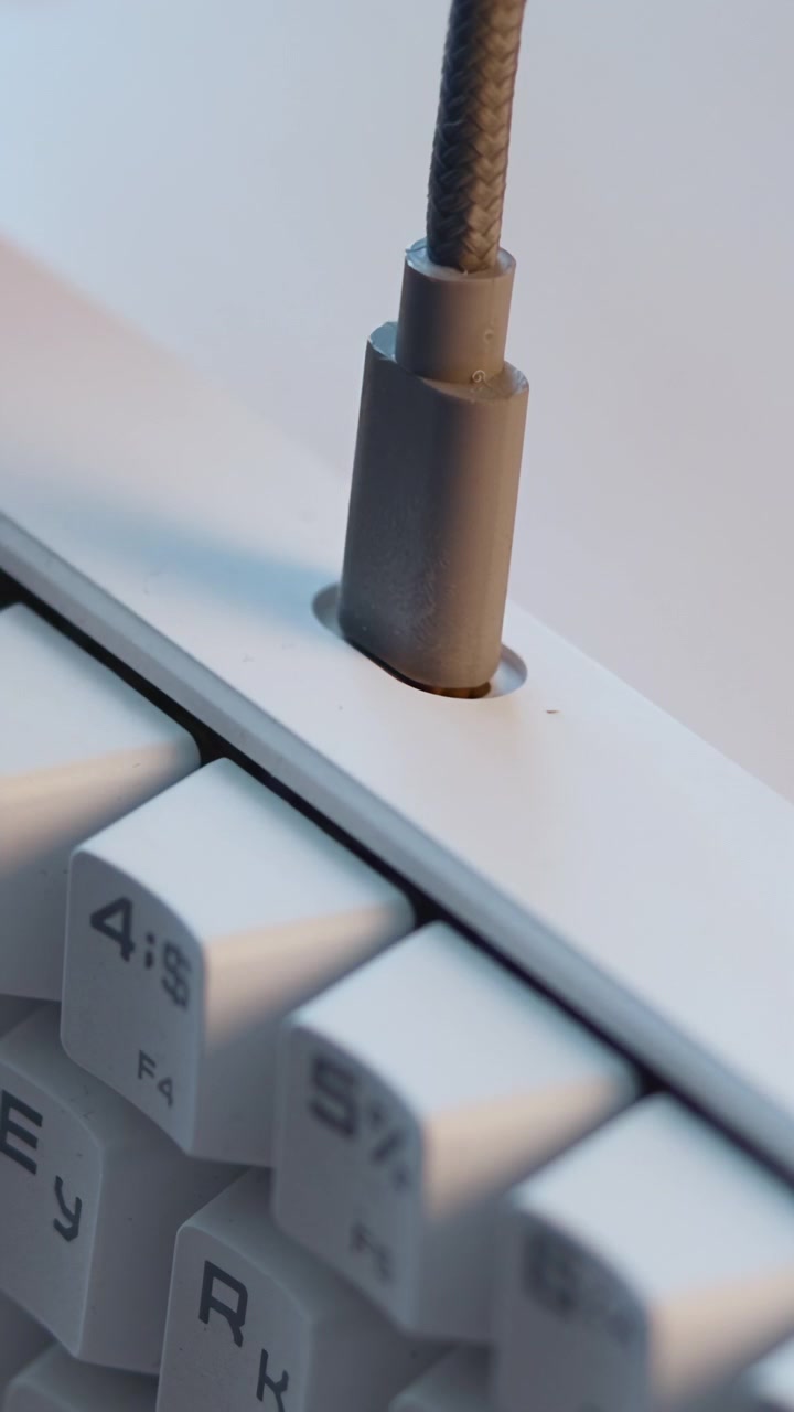 Near-macro low-angle view shows gray cable plug aligned with recessed rectangular port on white keyboard, then inserted into top edge, with soft lighting, shallow depth focus and clean workspace