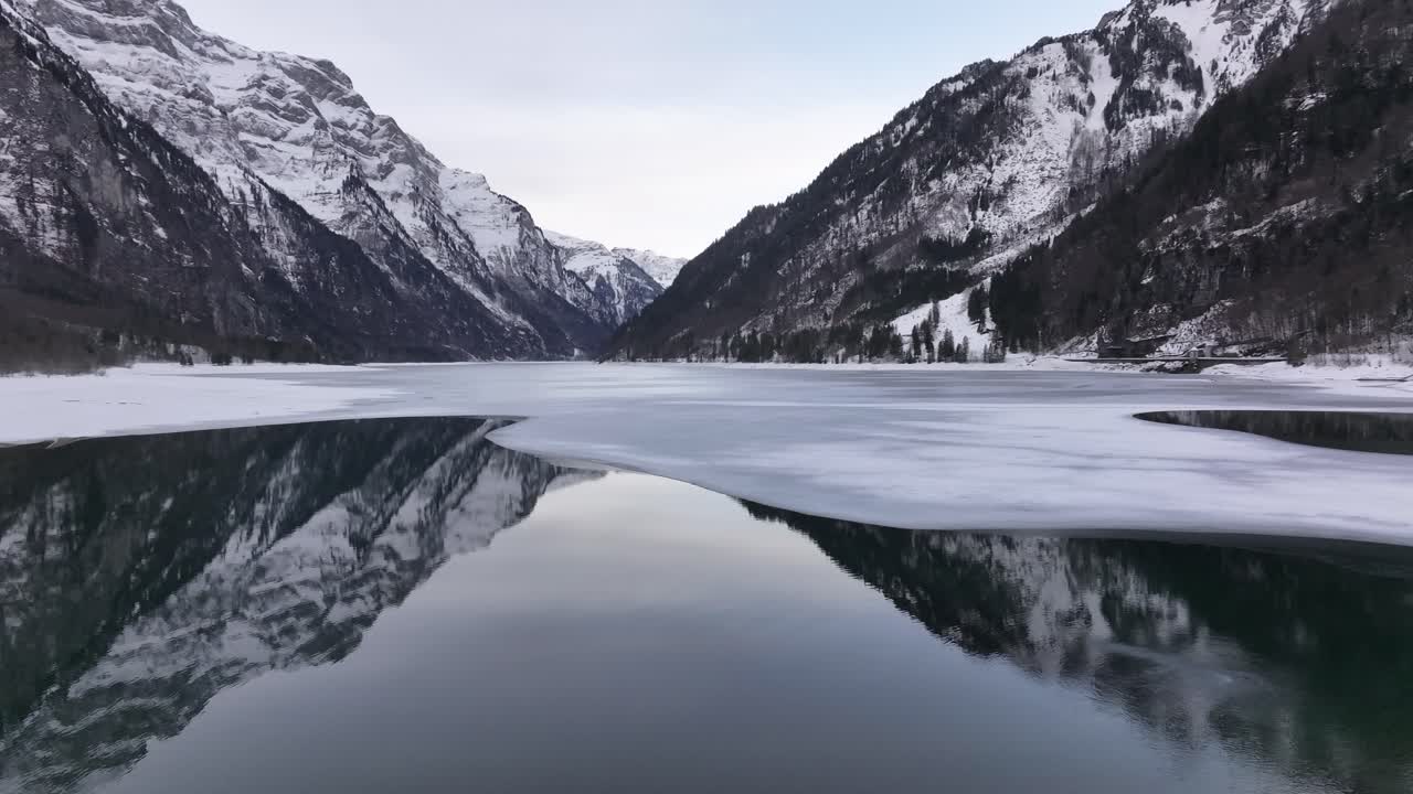 Partially frozen Klöntalersee lake with mirror-like waters reflecting the surrounding Swiss mountains, creating a serene and wintery landscape.