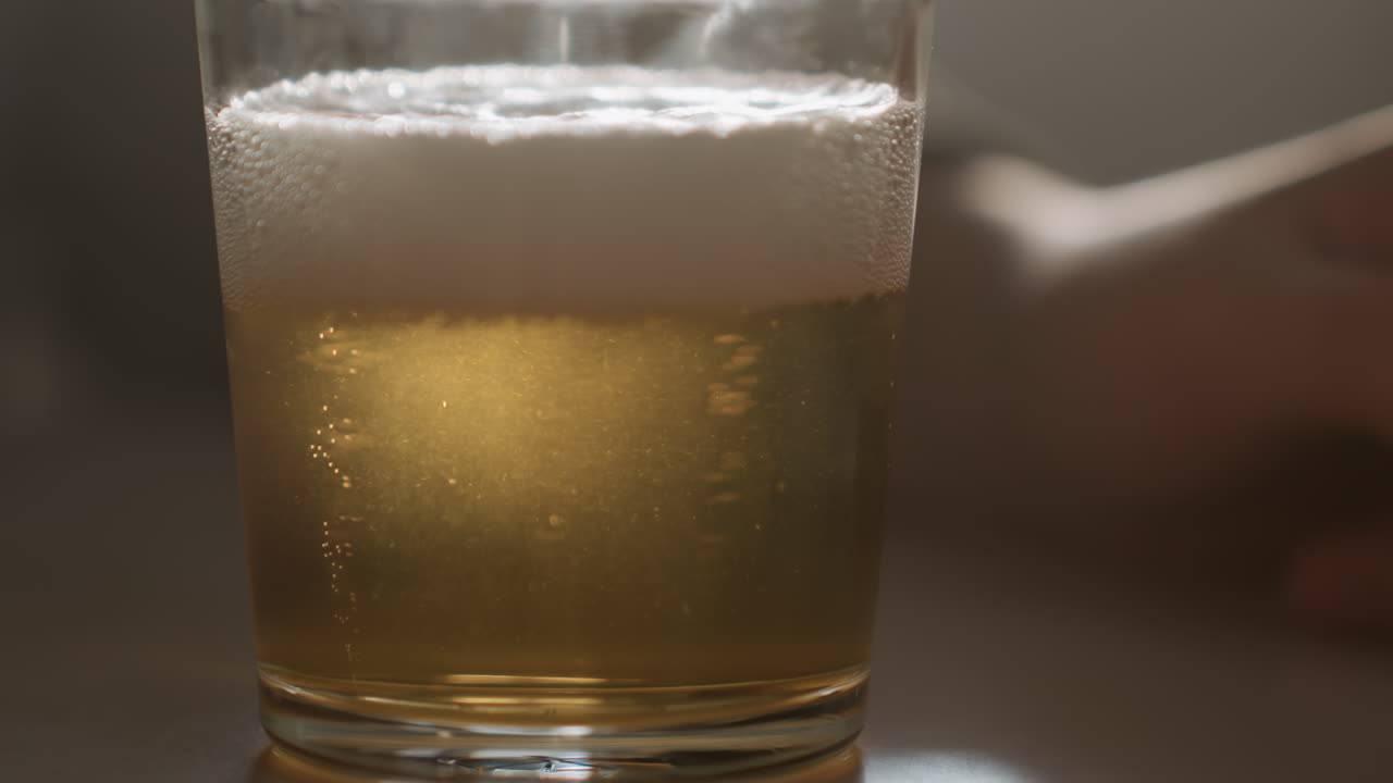 Primo piano di bicchiere di birra posato con schiuma, bollicine delicate che salgono in controluce caldo, braccio sfocato che riposa sullo sfondo, scena da tavolo accogliente e minimale con atmosfera serena e suggestiva, ideale per bevande.