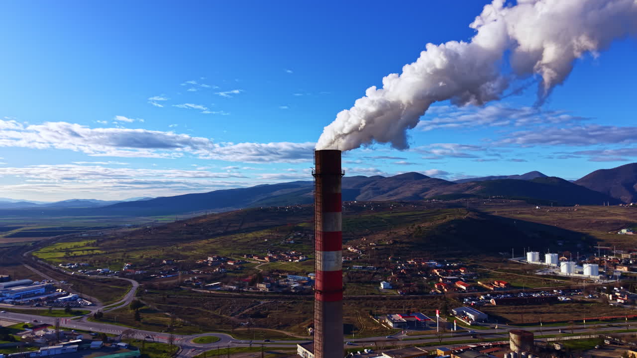 Il fumo si alza da un'alta ciminiera in un sito industriale vicino a una catena montuosa sotto un cielo azzurro e limpido nel pomeriggio.