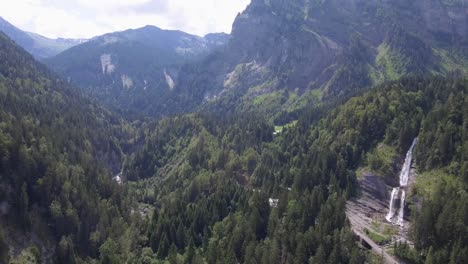 A-drone-flight-over-a-lush-green-alpine-forest-toward-a-cascading-glacier-melt-waterfall-tumbling-over-a-cliff-side