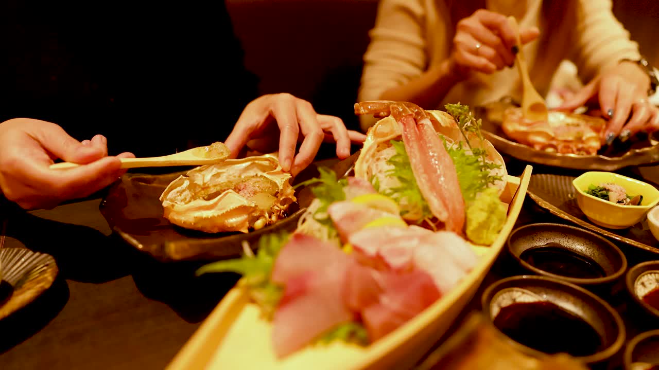 Primo piano di commensali che mangiano sashimi fresco e zampe di granchio da un piatto a forma di barca in legno.