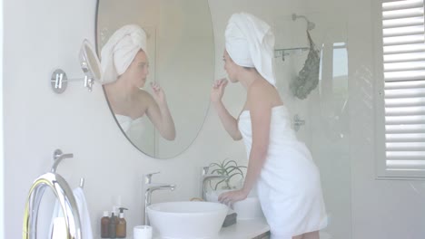 caucasian woman brushing teeth in front of mirror with towel on head in bathroom in slow motion