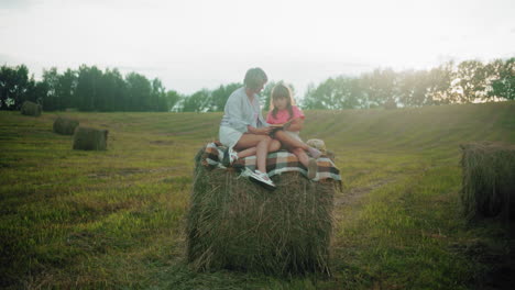 grandmother and granddaughter sit on hay bale reading novel in peaceful farmland surrounded by lush green fields, tranquil outdoor bonding moment with book, nature, and warm golden sunlight