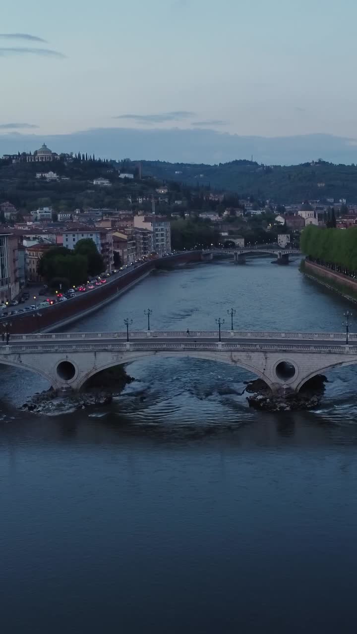 Vertical aerial shot of Verona showing Ponte della Vittoria bridge over the Adige River at blue hour, with hills, traffic, and buildings on both riverbanks, north of Italy, real time static camera