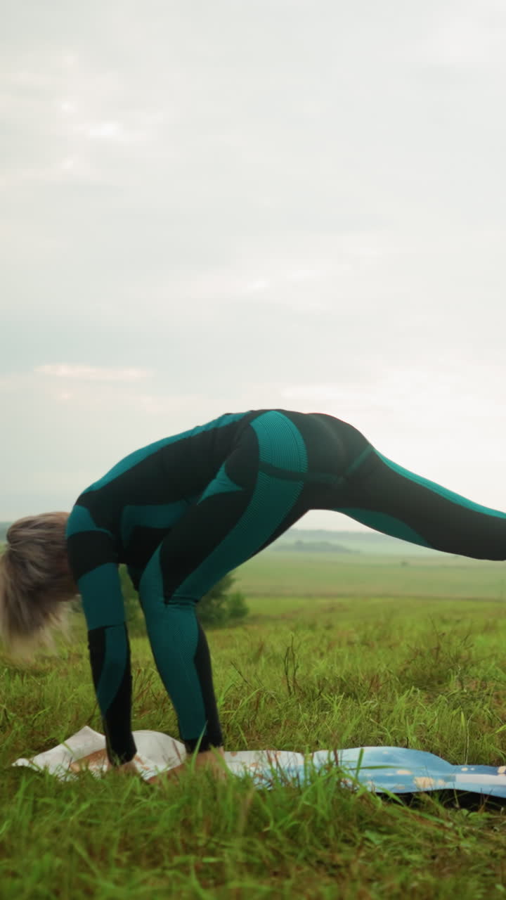 mujer al aire libre en un vasto campo de hierba en una alfombra de yoga practicando una postura baja, inclinándose hacia adelante con sus manos tocando la alfombra, bajo un cielo nublado, rodeada de árboles en el horizonte
