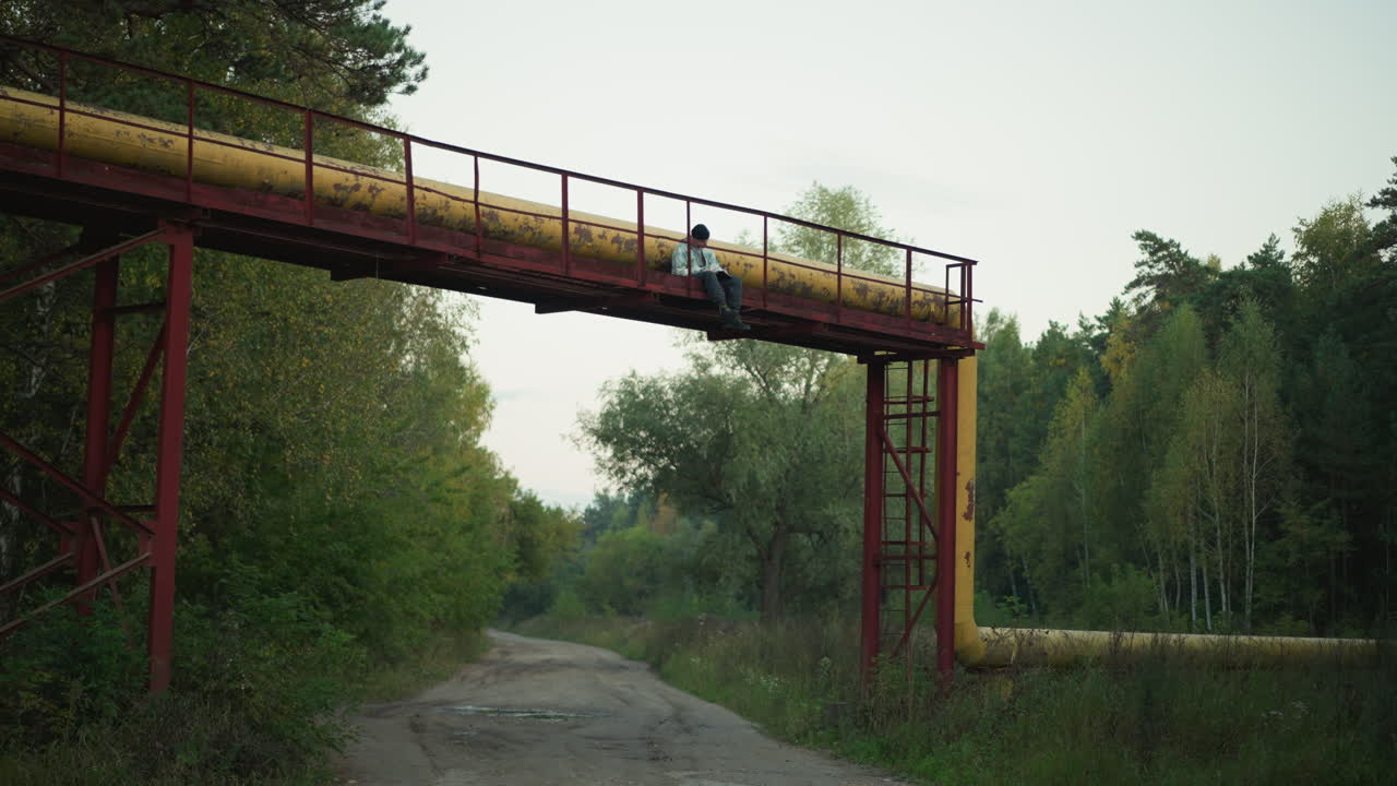 Rusten rørledningsbro over skovvej, enlig vandrer stopper midt på broen, grusvej nedenunder, omkringliggende birk og fyr, blødt aftenlys, dæmpet grøn palet, rolig udforskningsstemning, rejse