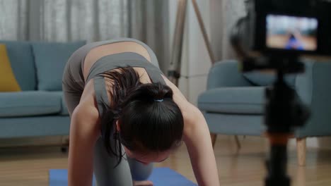 young asian trainer female in sports clothes speaking to camera and doing yoga in forearm plank pose while recording teaching exercise at home