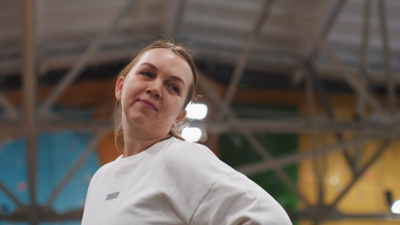 White woman badminton coach in indoor court scanning practice, confident posture with subtle smile, steel truss ceiling and colorful banners visible, neck turn and profile shots convey leadership