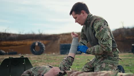 seitenansicht eines selbstbewussten männlichen militärs in einer grünen tarnuniform mit medizinischen handschuhen, der die hand mit stoffverbandungen eines verwundeten soldaten während einer trainingsübung auf einem militärischen trainingsplatz mit einer barrikade aus radreifen verbindet