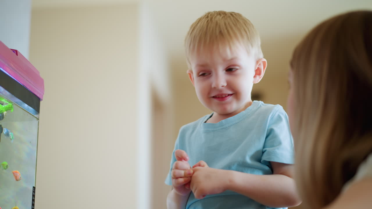 Young boy in blue shirt points at aquarium while sucking finger, observing colorful fish swimming among green plants inside glass tank, showing concentration, wonder and childhood curiosity in calm indoor