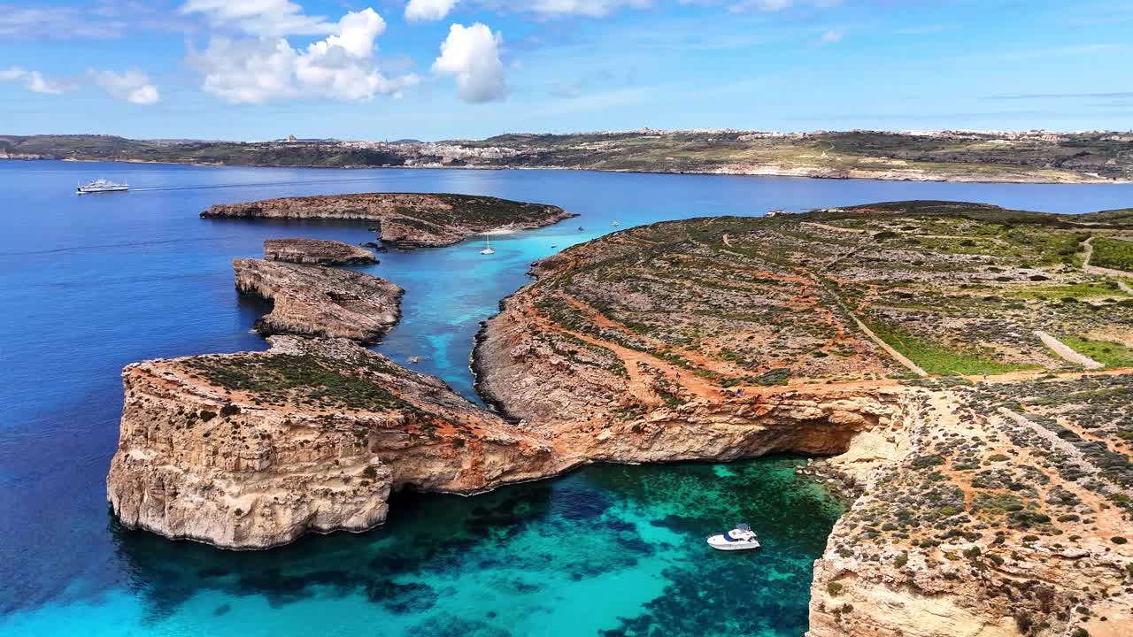 Vista aerea della Blue Lagoon a Comino, Malta, che mostra una baia rocciosa, piccoli isolotti, barche ancorate e un mare turchese cristallino sotto la luce del sole.