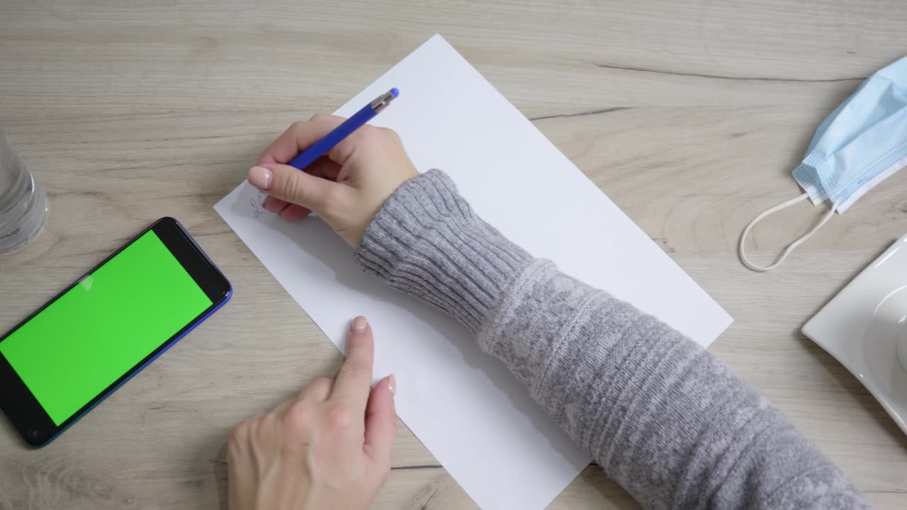 Conference call on a phone with a green screen, work from home. Shot from above, female hands writing on paper. Work at the time of the Covid19 pandemic, there is a mask on the table.