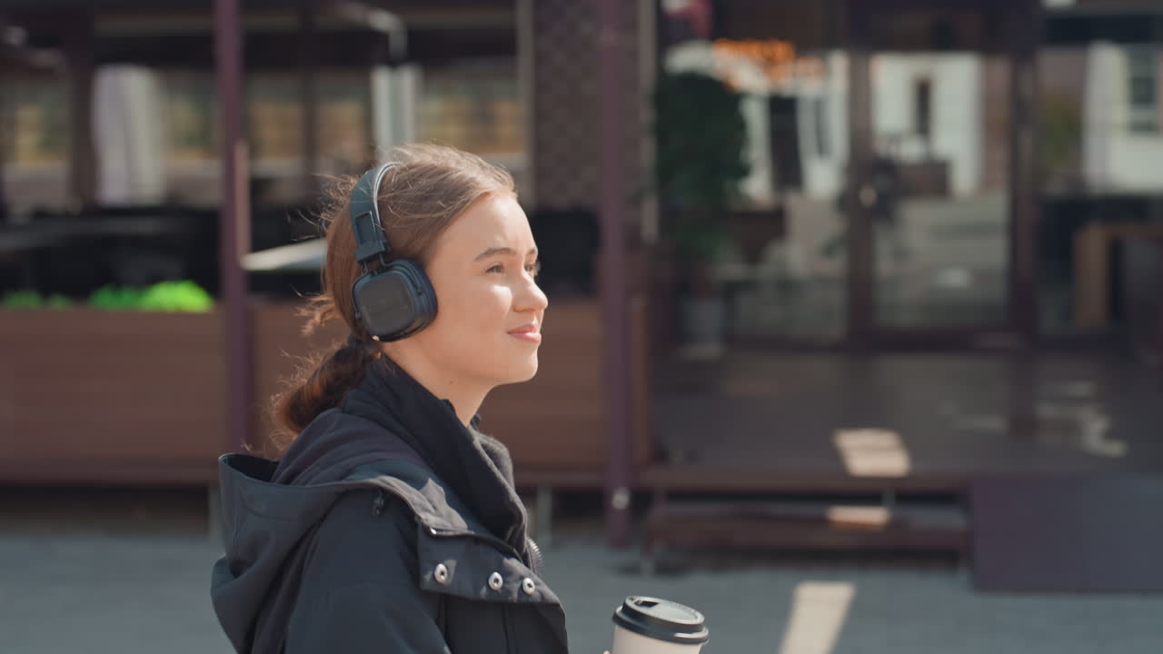 Donna caucasica che cammina con un caffè, indossa cuffie e una giacca invernale, passa davanti al dehor di un bar e a delle aiuole, sorseggia una tazza da asporto, espressione pensierosa, soffice luce del sole mattutino, marciapiede urbano, lentamente