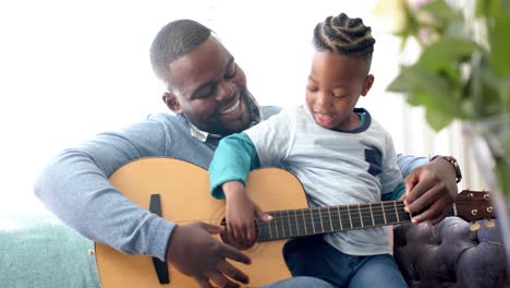 happy african american father and son sitting on sofa and playing guitar, in slow motion