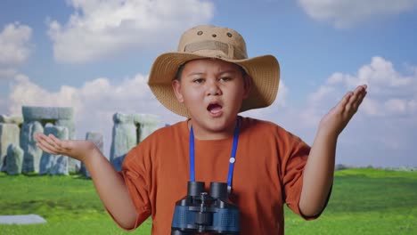 asian boy with a hat standing doubtfully and asking why after looking through the binoculars. boy researcher examines something while traveling in stonehenge, travel adventure concept, close up