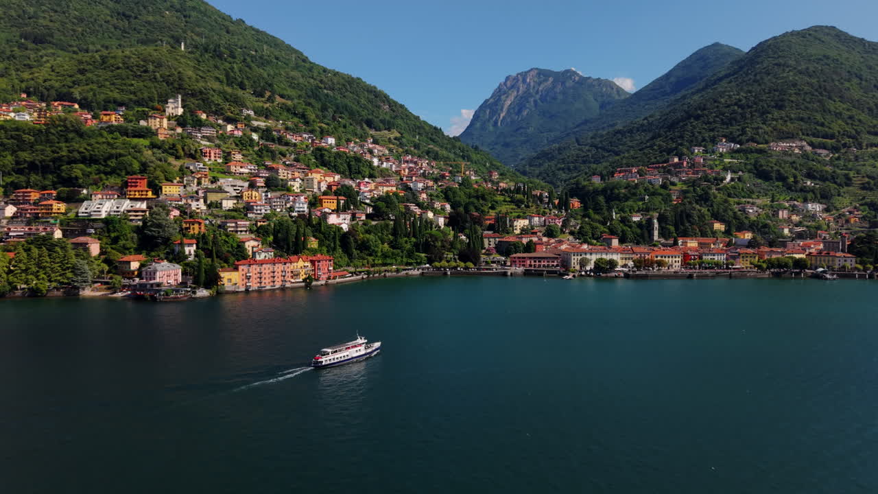 Drone orbits a ferry on Lake Como, rising to reveal the Italian lakeside village and surrounding green mountains under clear daylight