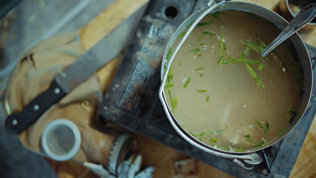 Overhead view of cooking pot filled with broth and herbs on old stove, placed next to worn gas mask and knife on wooden table, symbolizing survival, resilience, and harsh living conditions indoors