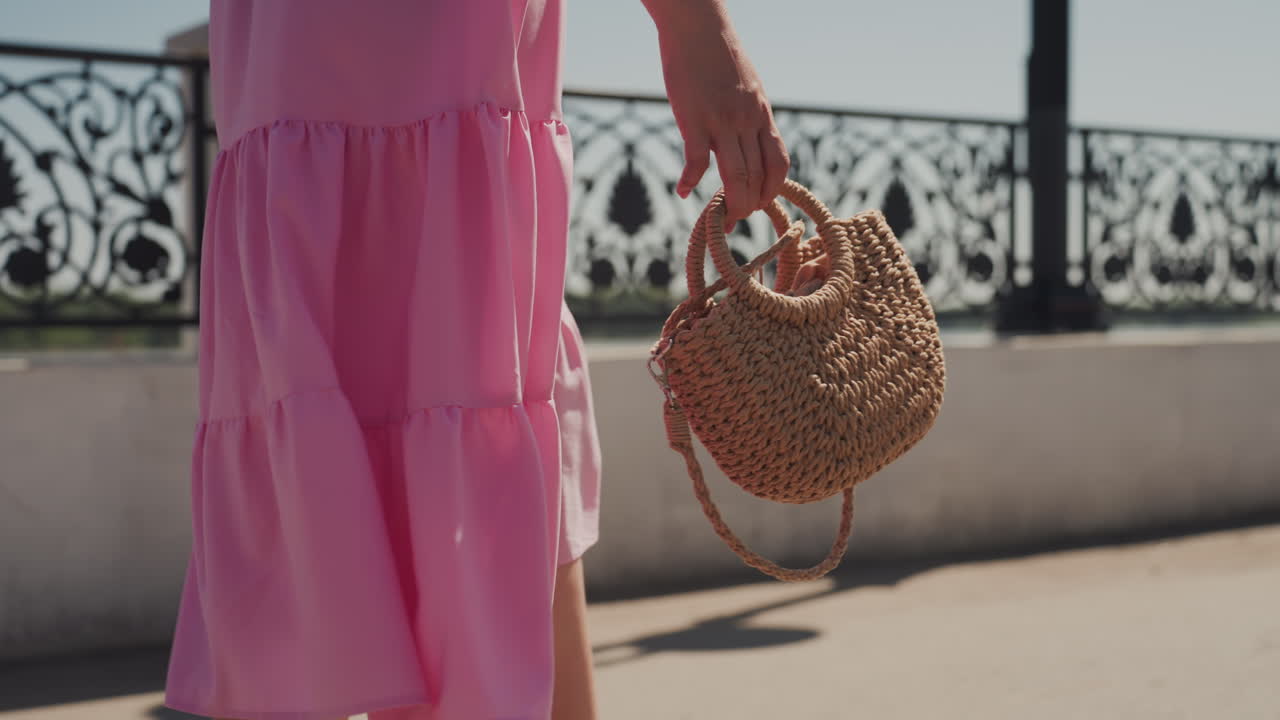 Abito prendisole rosa abbinato a una borsa di paglia mentre cammina sul lungomare illuminato dal sole accanto a un'ornata ringhiera in ferro, gonna a balze che ondeggia al vento mentre la modella avanza, borsa in tessuto bohémien che oscilla, atmosfera calda e cinematografica
