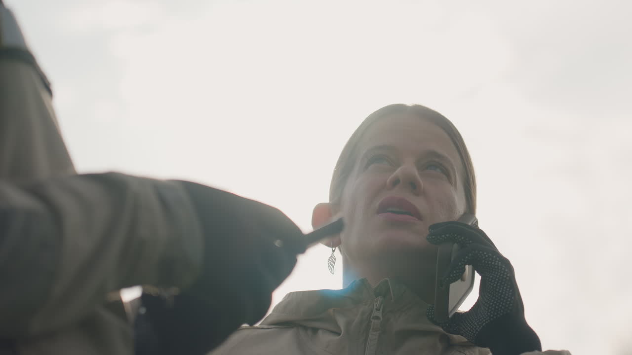 woman on phone observing sky with sunlight beam behind turning head while colleague stands nearby in grassy field monitoring aerial sighting device instructions relayed via urgent call