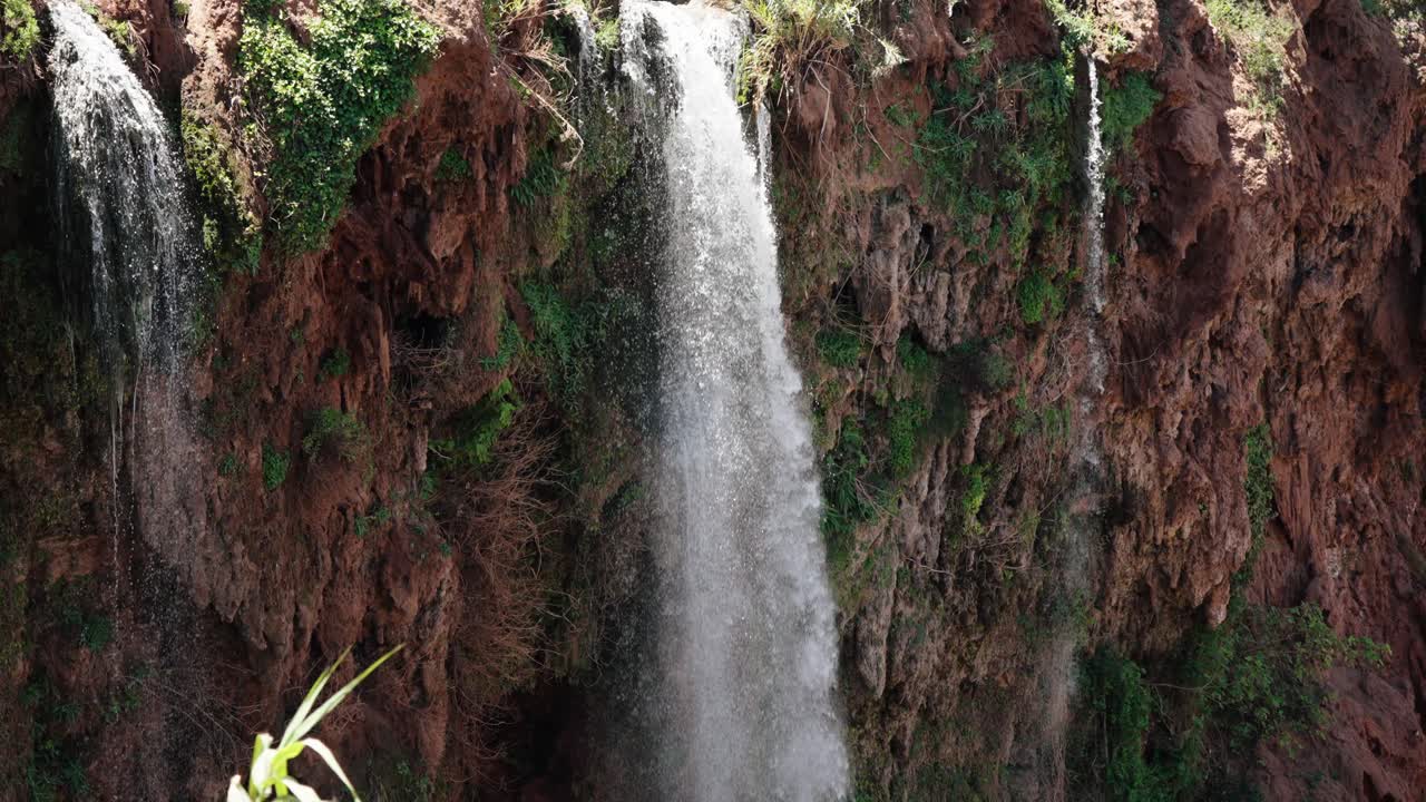 Slow-motion view of upper Ouzoud Waterfalls cascading down mossy cliffs with scattered green vegetation.