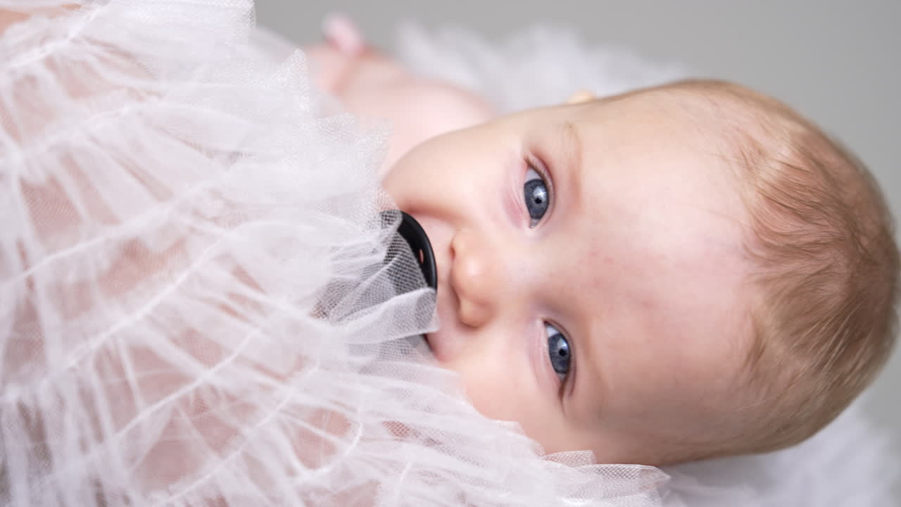 Face of an adorable grey-eyed Caucasian baby with pacifier. Infant swayed in mom's hands falling asleep. Close up.