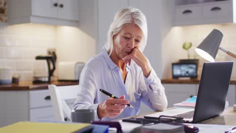 thoughtful senior caucasian woman sitting at table in kitchen, using laptop