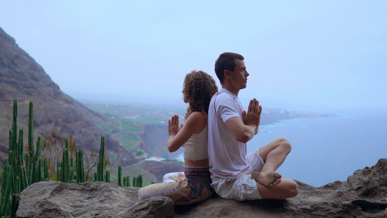 On a rock at the mountain's crest, a man and woman meditate and practice yoga back to back, gazing out at the ocean's expanse