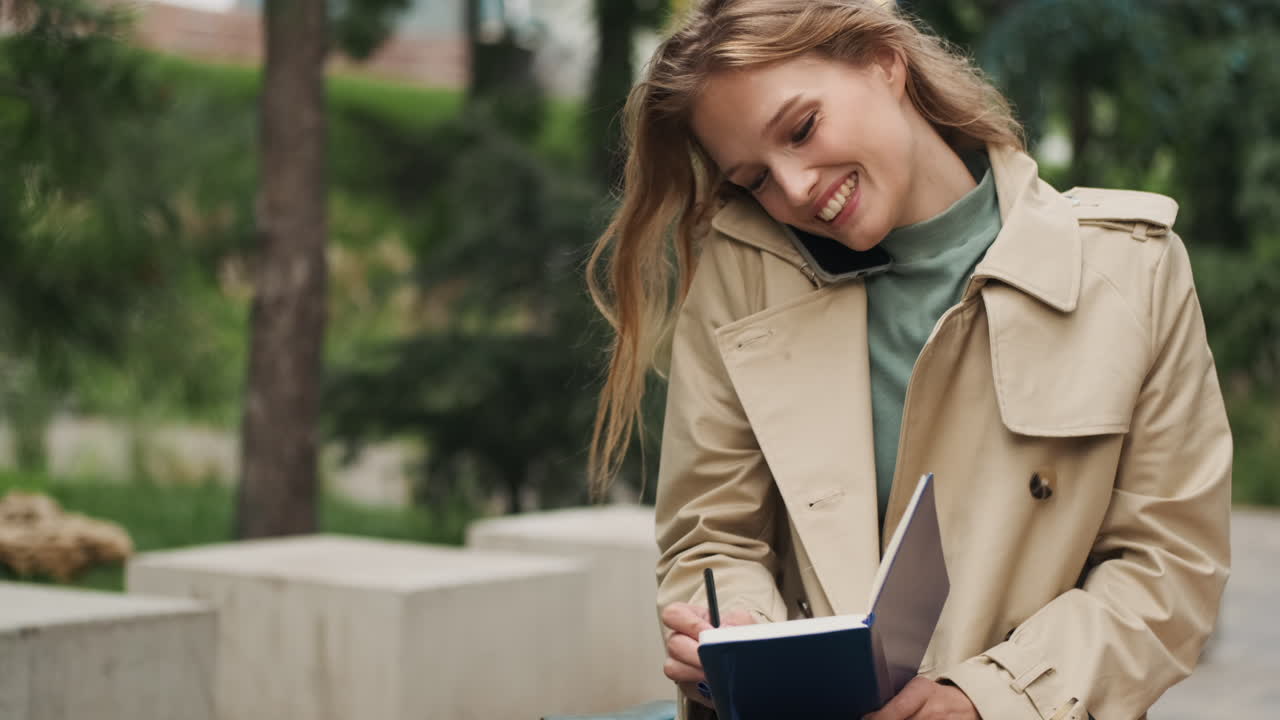 Caucasian female student talking on the phone while writing in a notebook outdoors.