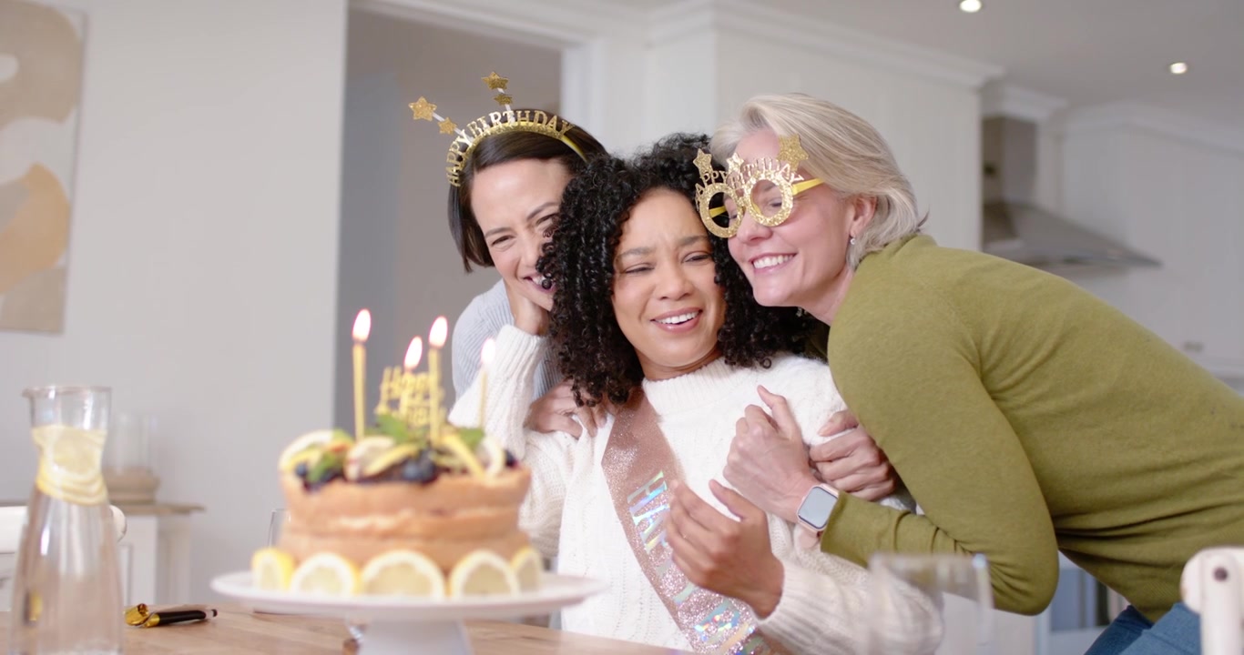 Amigas diversas incentivando aniversariante a soprar velas acesas em bolo na mesa da cozinha
