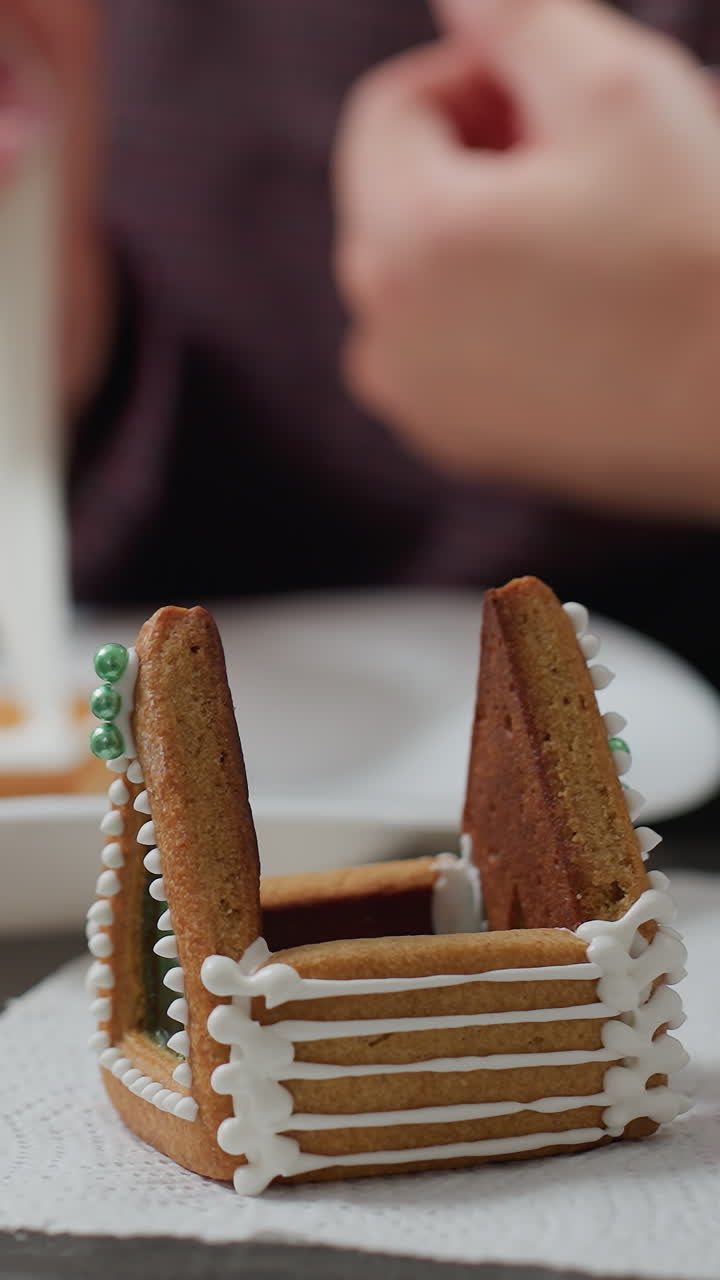 구슬로 된 팔를 가진 여성의 흐릿한 시선 조심스럽게 접시에 아이싱 쿠키, 주방 설정에서 조직에 배치 된  아이싱과 녹색 식용 진주와 함께 장식 된 진저브레드 집