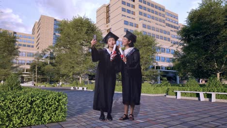 cuerpo lleno de estudiantes asiáticas graduadas con gorras y vestidos con diplomas tomando fotos en el teléfono inteligente mientras están de pie frente a un magnífico edificio universitario