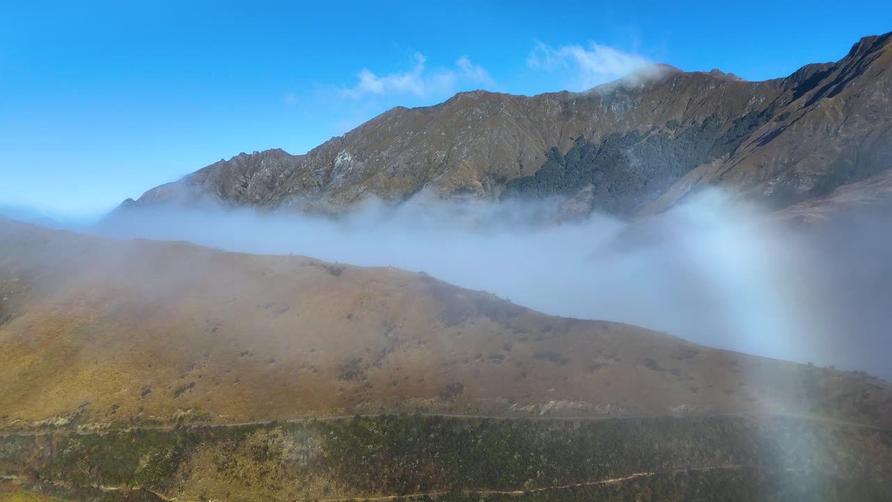 Aerial view of mist rolling over mountains near Moke Lake, Queenstown. Clear blue sky contrasts with the foggy landscape
