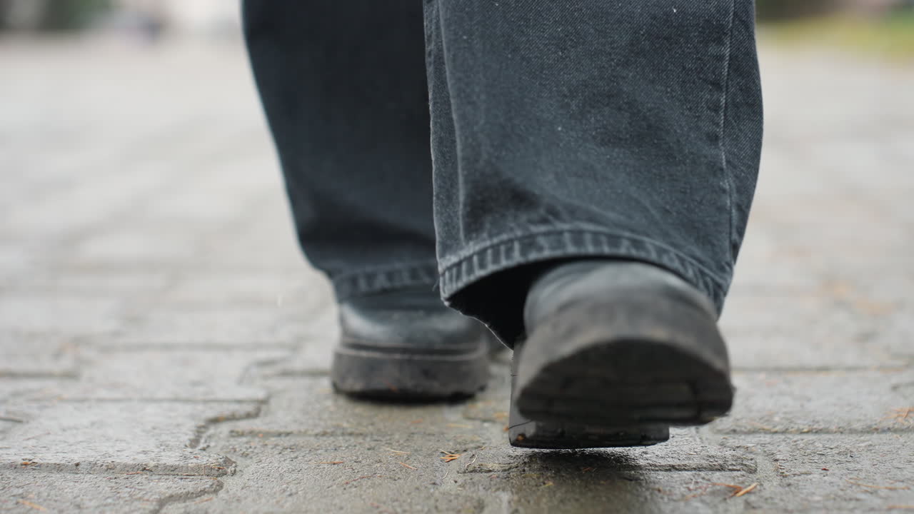 Close up of someone's black trousers and black boots walking slowly forward on outdoor paved path during calm overcast day, focusing on footwear movement, step rhythm, and cool atmospheric